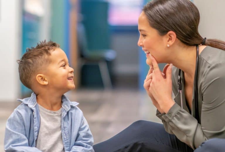 Speech therapy session female speech therapist smiling at a small child and pointing at the corners of her mouth as the the child watches and smiles back
