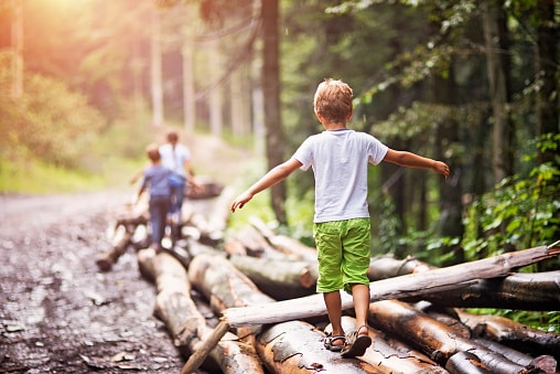 Children Pediatric Balance Disorder A child is walking on a pile of downed logs on the edge of a fire road. His arms are spread out for balance. His parents are 50 feet ahead of him.