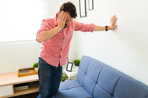 Man Experiencing Dizzy Spell Man in pink shirt in an office setting holding his head with one hand while propping himself up against a wall with his other arm outstretched.