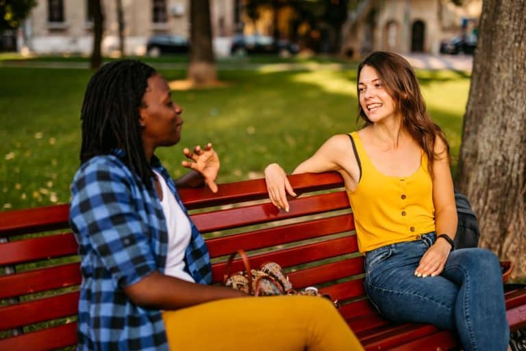 speech-therapy-aphasia-scaled two women talking on a red park bench, one is black and wearing a blue shirt and yellow pants, the other is white and wearing a yellow shirt and blue pants