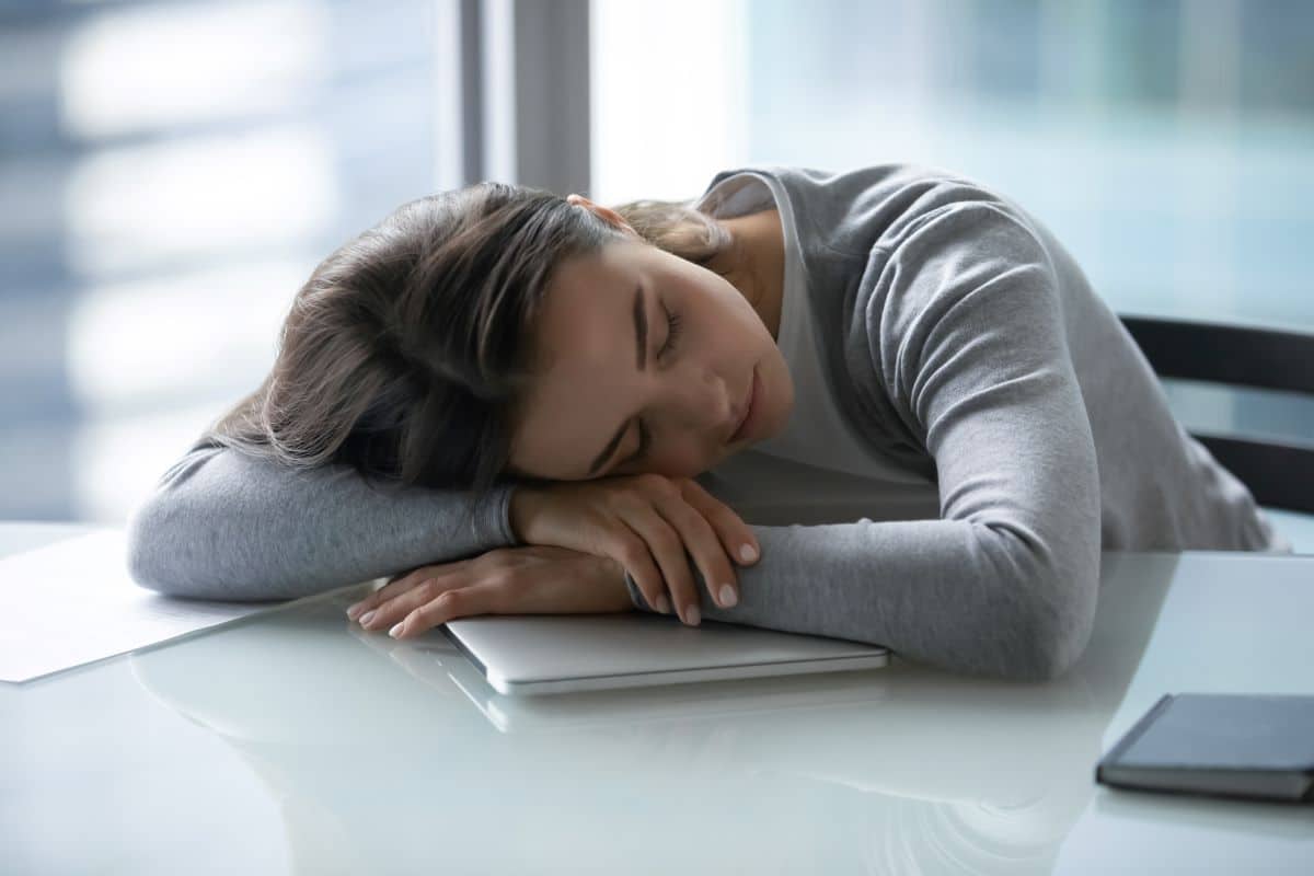 Sleep-Narcolepsy Young woman who has fallen asleep on an office desk with a closed laptop underneath her. She is resting her head on her crossed arms.